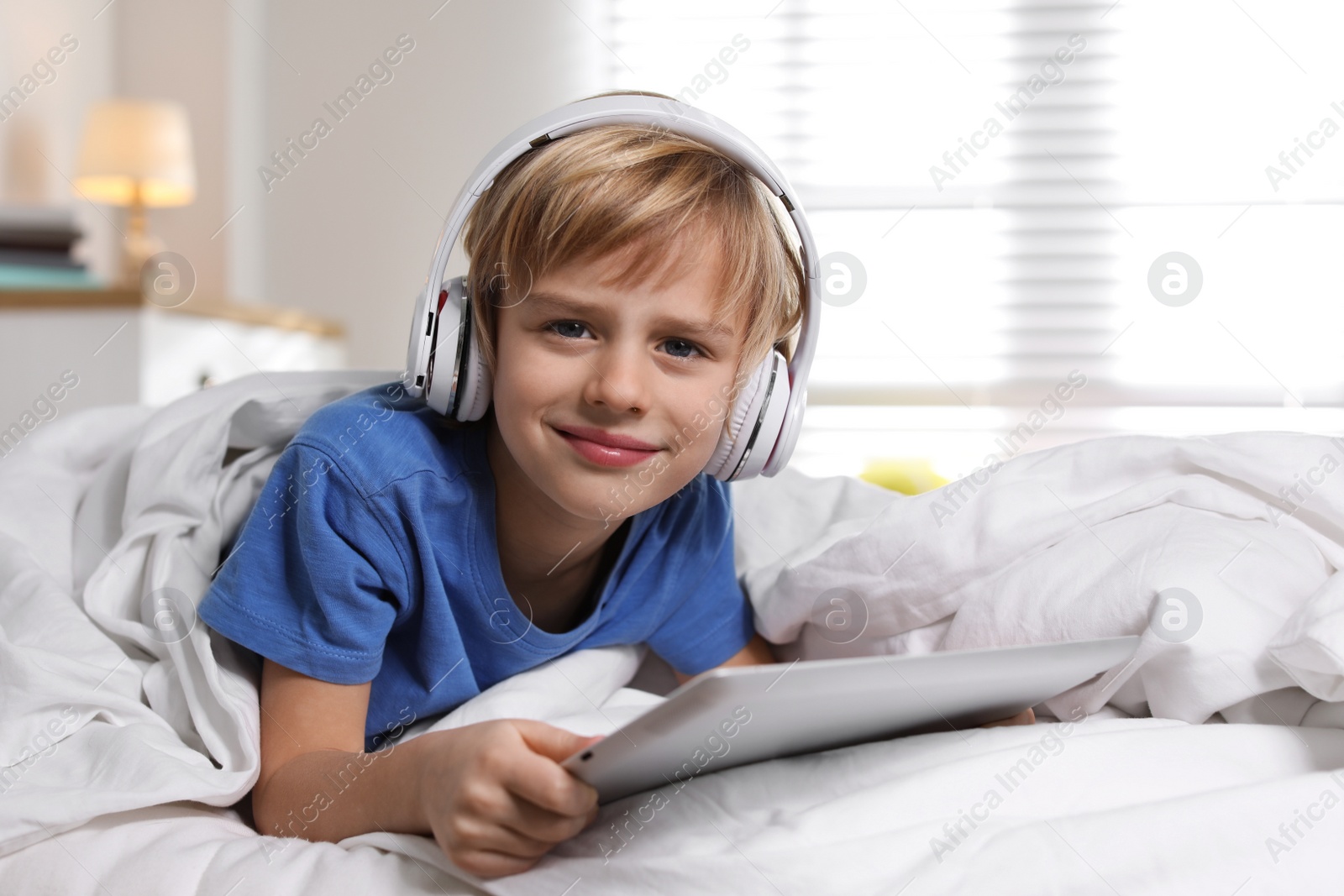 Photo of Cute little boy with headphones and tablet listening to audiobook in bed at home