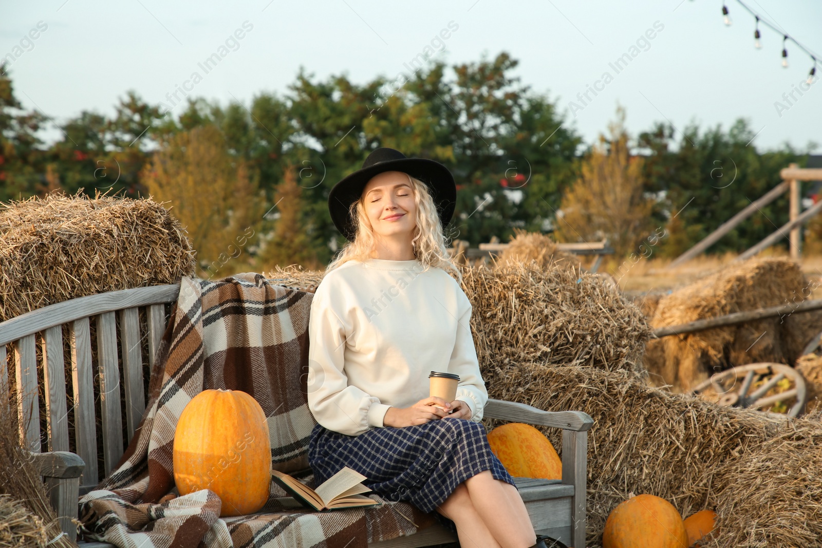 Photo of Beautiful woman with cup of hot drink sitting on wooden bench near hay bales and pumpkins outdoors. Autumn season