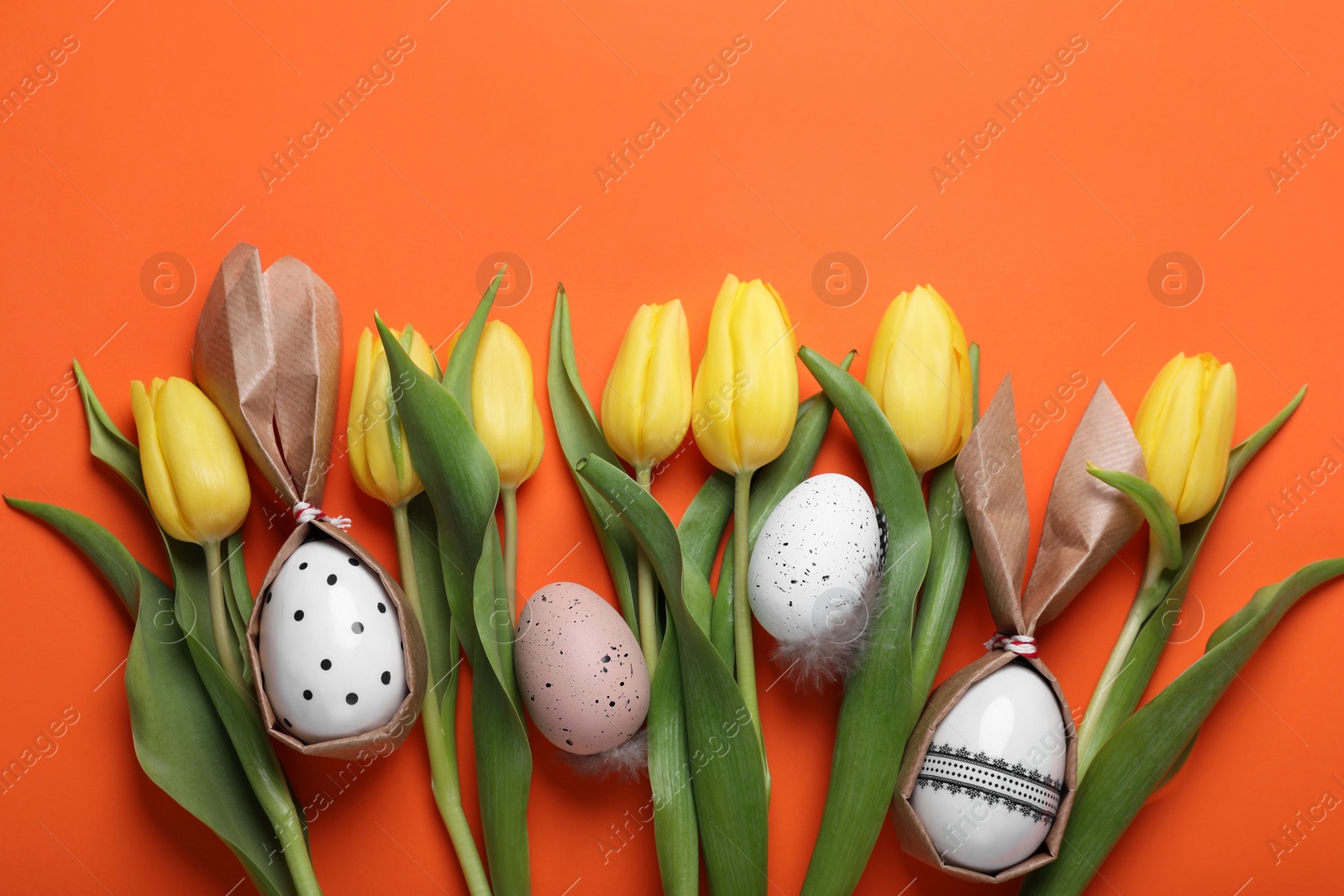 Photo of Easter bunnies made of craft paper and eggs among beautiful tulips on orange background, flat lay