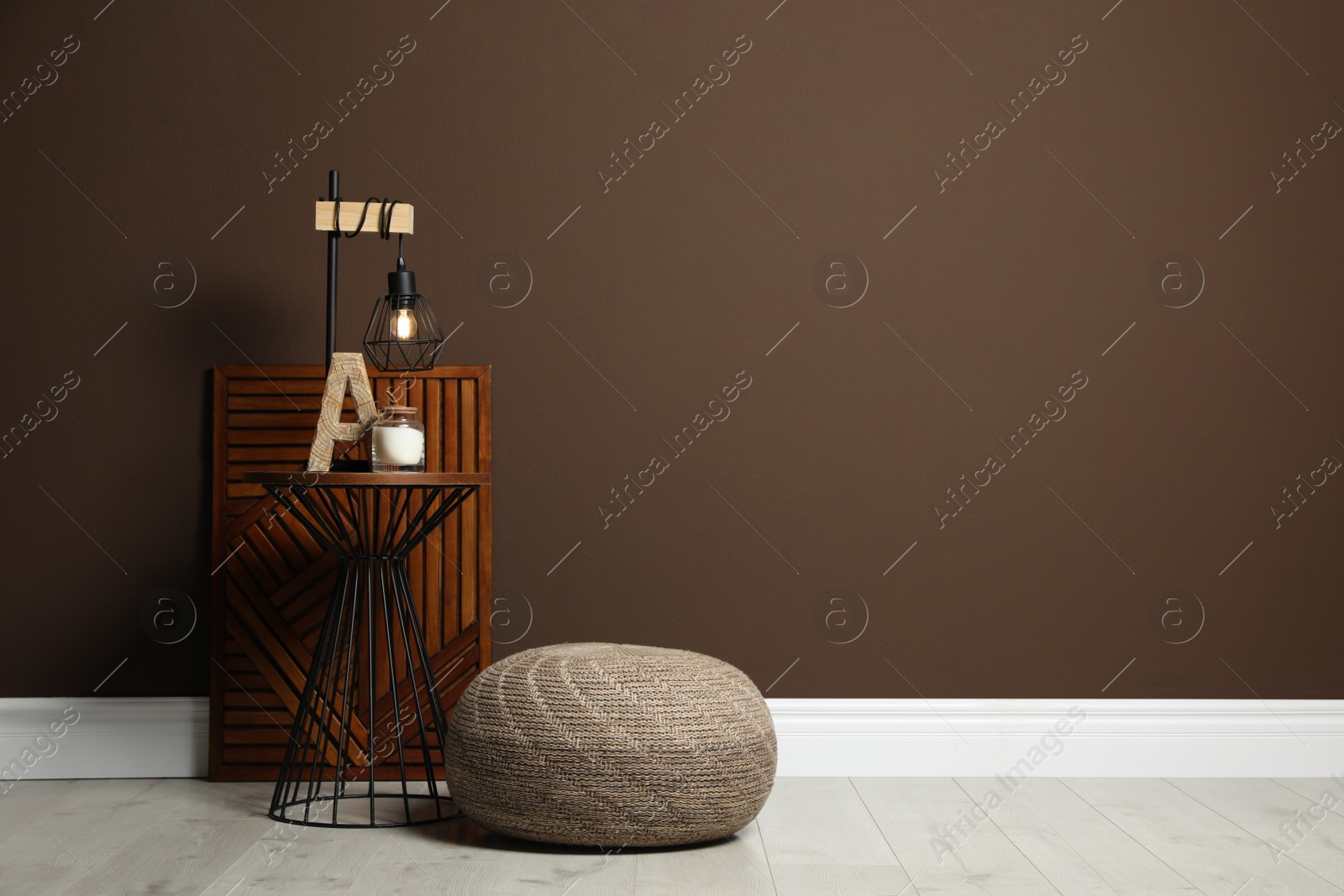 Photo of Comfortable knitted pouf and table with decor elements near brown wall indoors, space for text. Interior design