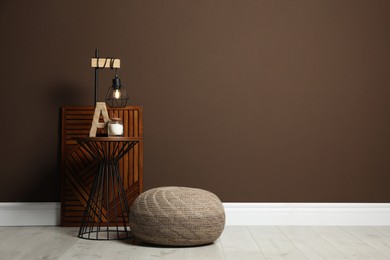 Photo of Comfortable knitted pouf and table with decor elements near brown wall indoors, space for text. Interior design