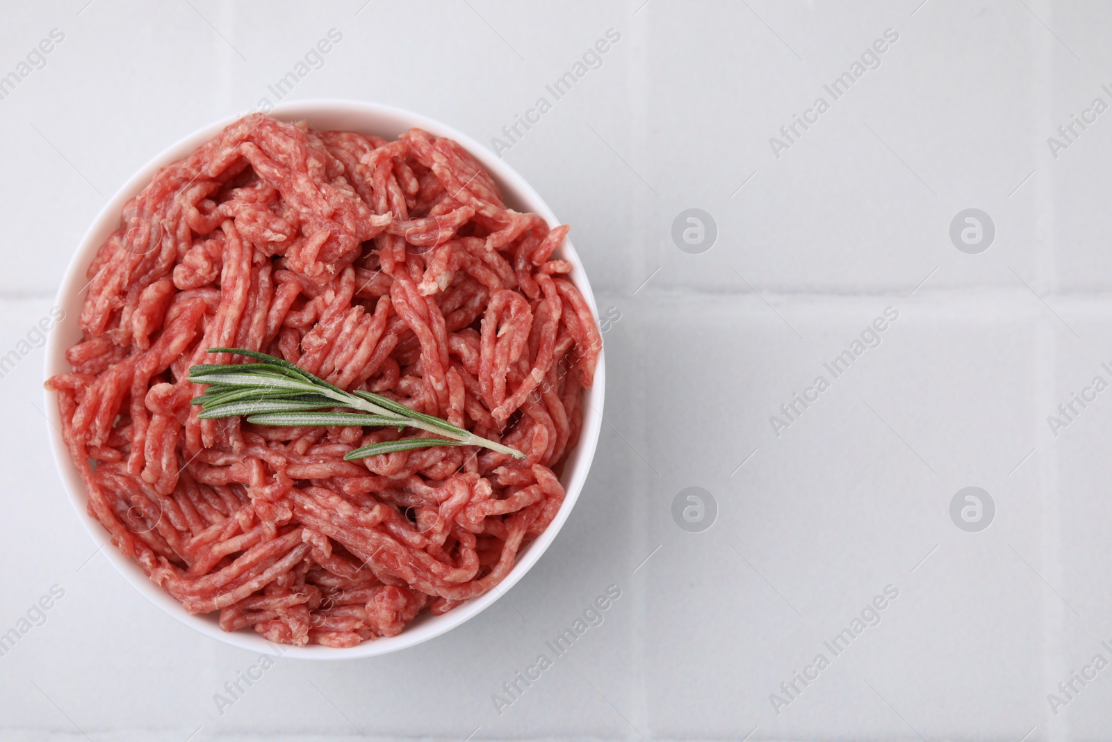 Photo of Fresh raw ground meat and rosemary in bowl on white tiled table, top view. Space for text