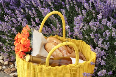 Photo of Yellow wicker bag with beautiful roses, bottle of wine and baguettes near lavender flowers outdoors