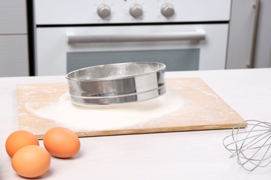 Photo of Sieve with flour, eggs, whisk and rolling pin on table in kitchen