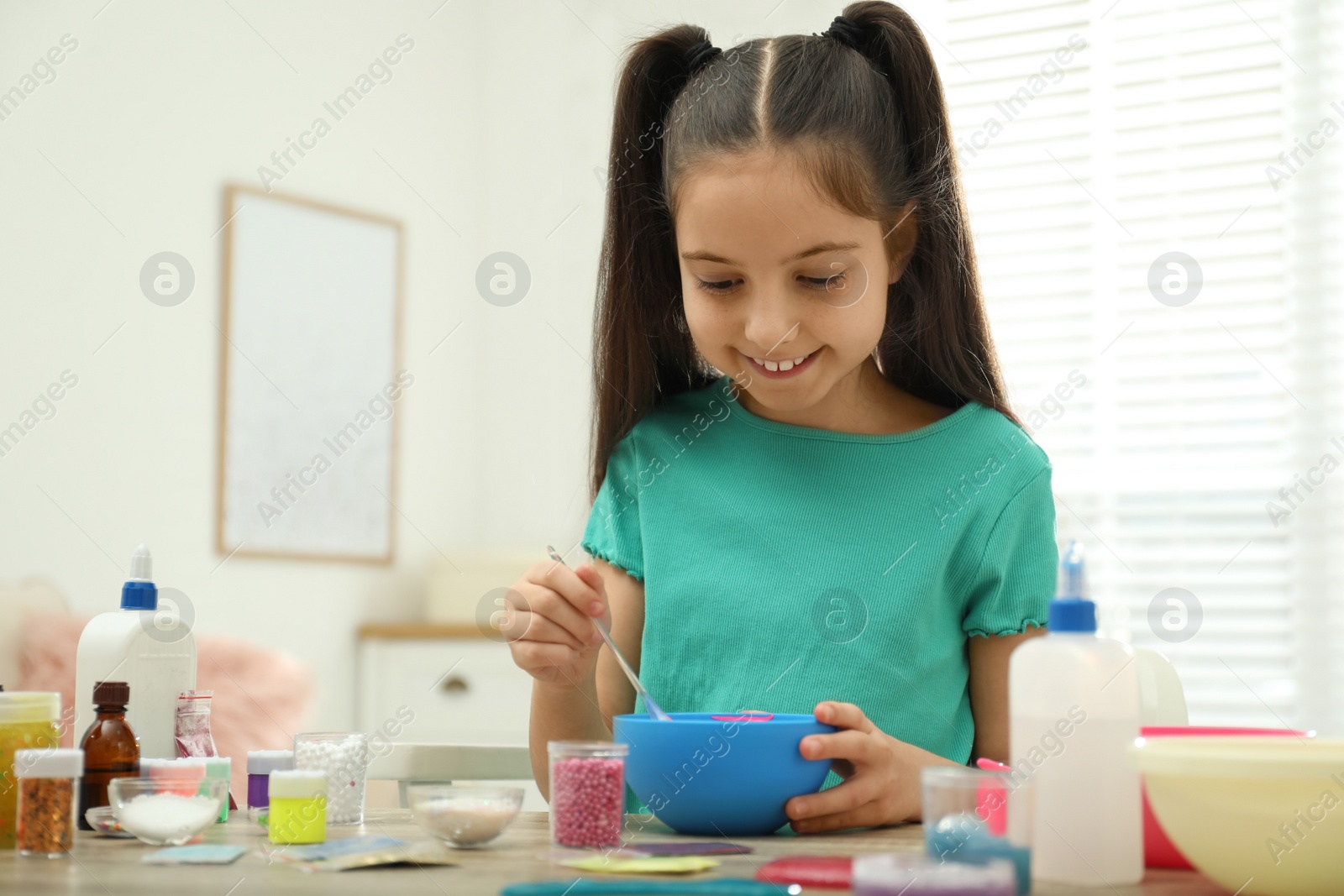 Photo of Cute little girl mixing ingredients with silicone spatula at table in room. DIY slime toy