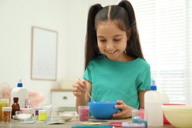 Photo of Cute little girl mixing ingredients with silicone spatula at table in room. DIY slime toy