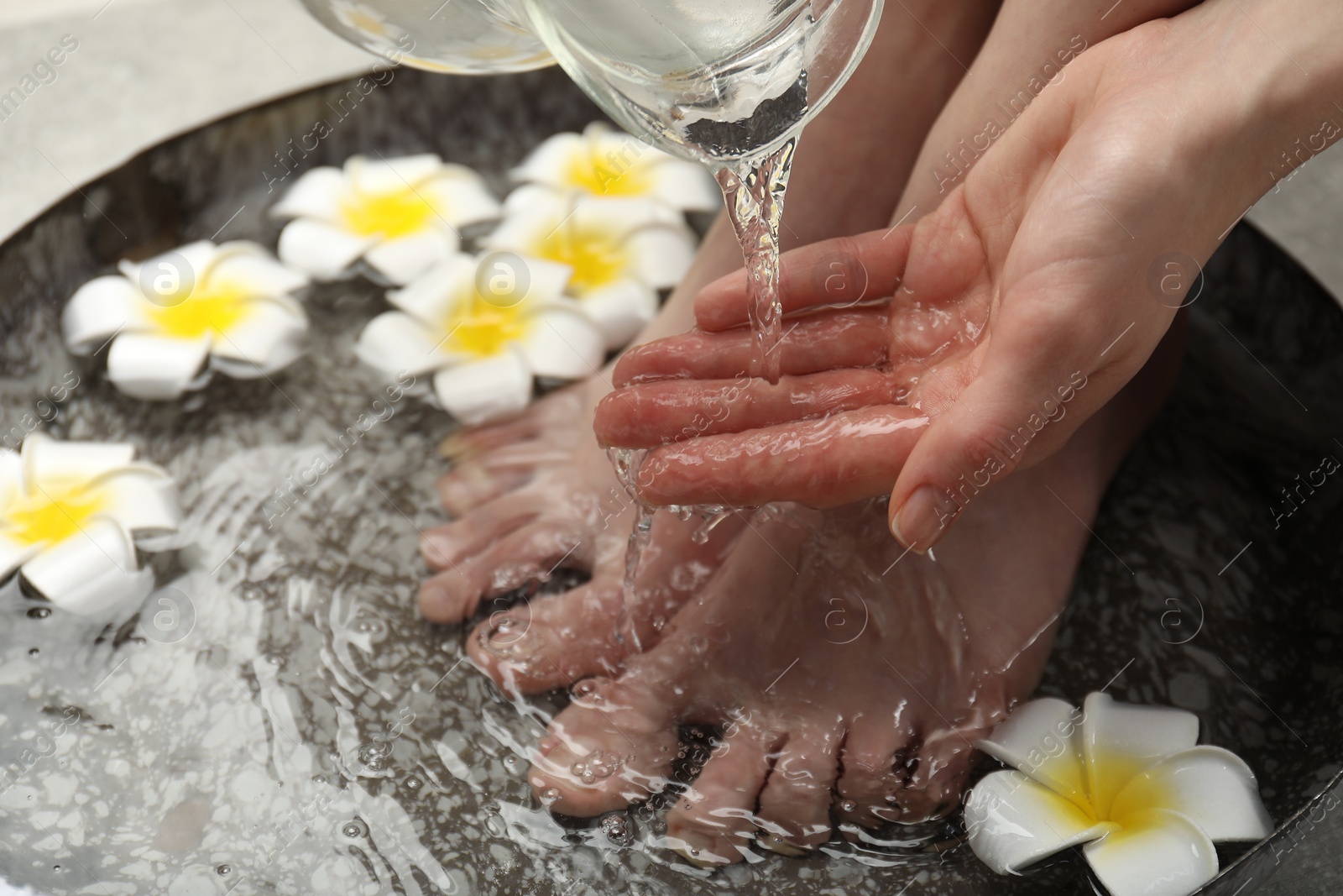 Photo of Woman pouring water onto hand while soaking her feet in bowl, closeup. Spa treatment