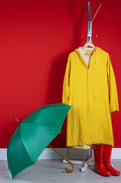 Photo of Stylish umbrella, raincoat and boots near red wall