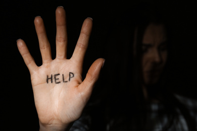 Abused young woman showing palm with word HELP against black background, focus on hand. Domestic violence concept