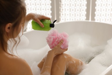 Woman pouring shower gel onto mesh pouf in bath indoors