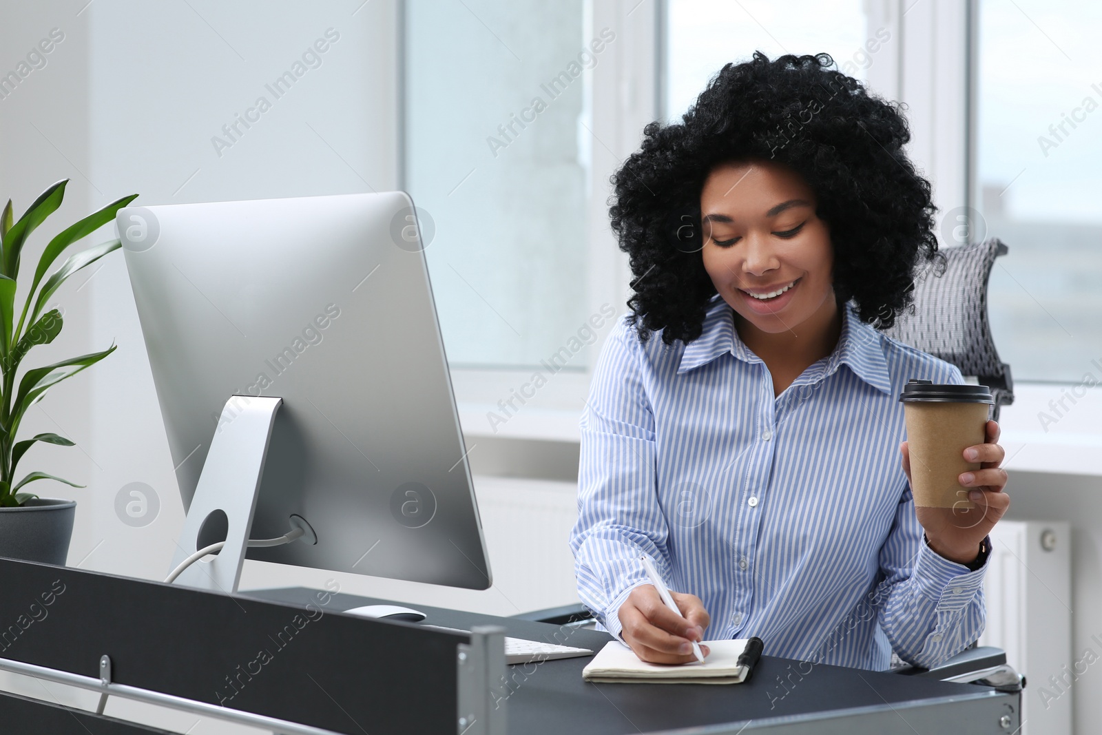 Photo of Young woman with cup of drink taking notes near computer at table in office