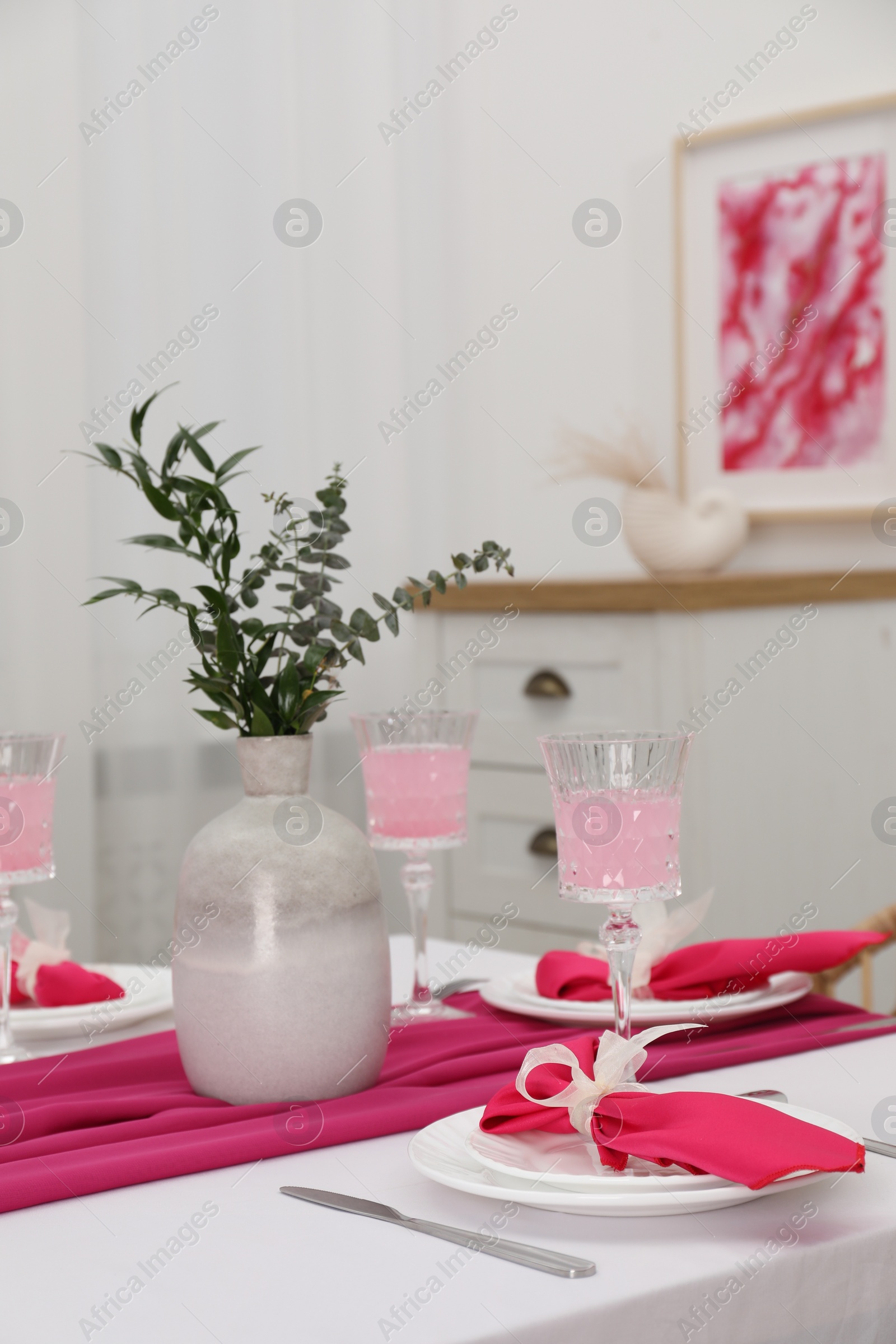 Photo of Table setting. Glasses of tasty beverage, plates, pink napkins and vase with green branches in dining room