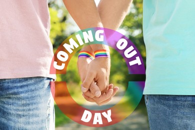 Image of National Coming Out day. Gay couple with pride rainbow wristbands holding hands outdoors, closeup