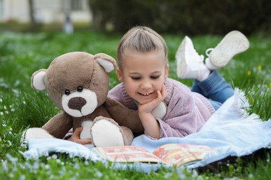 Photo of Little girl with teddy bear and book on plaid outdoors