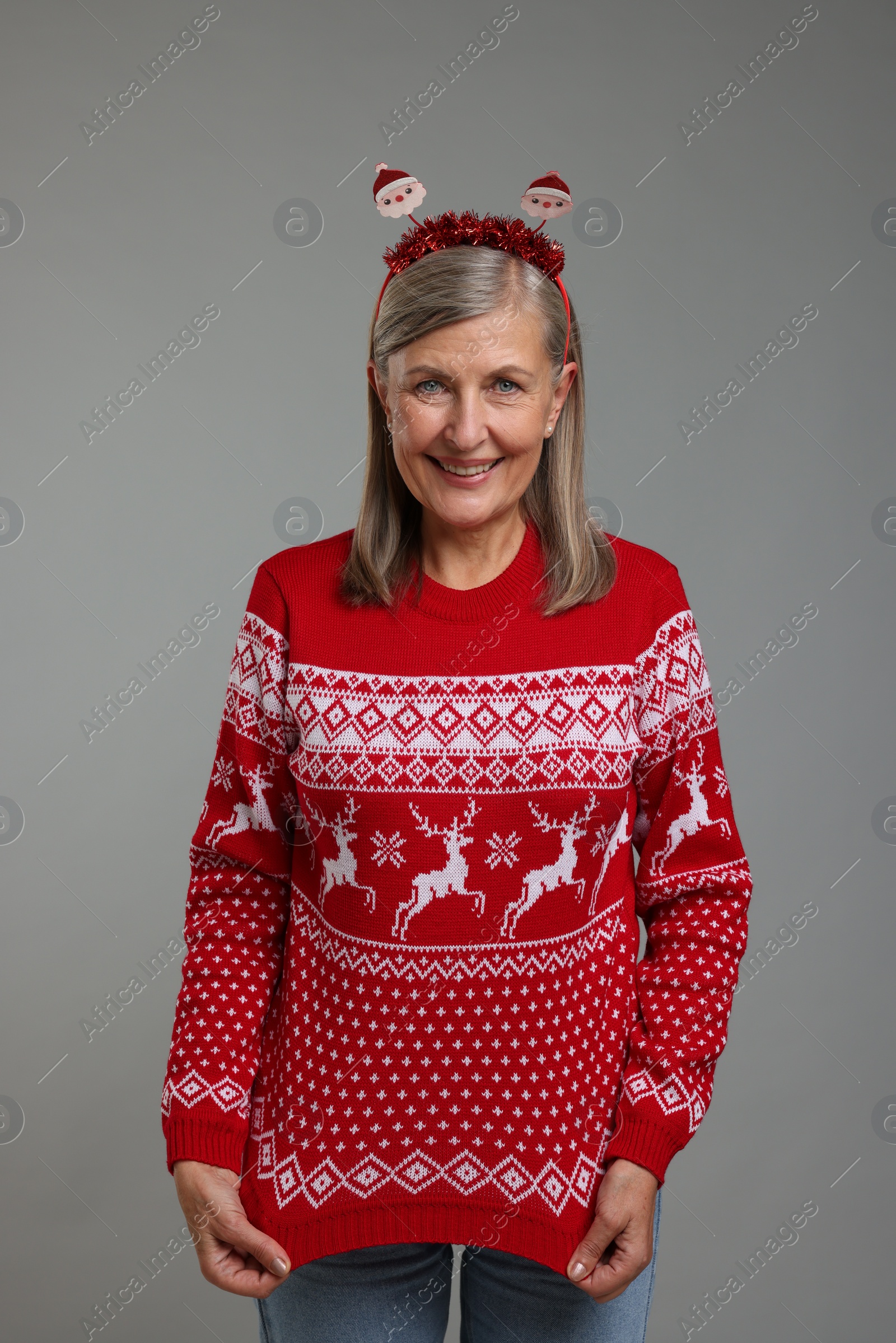 Photo of Happy senior woman in Santa headband showing her Christmas sweater on grey background