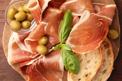 Photo of Slices of tasty cured ham, olives, bread and basil on wooden board, top view