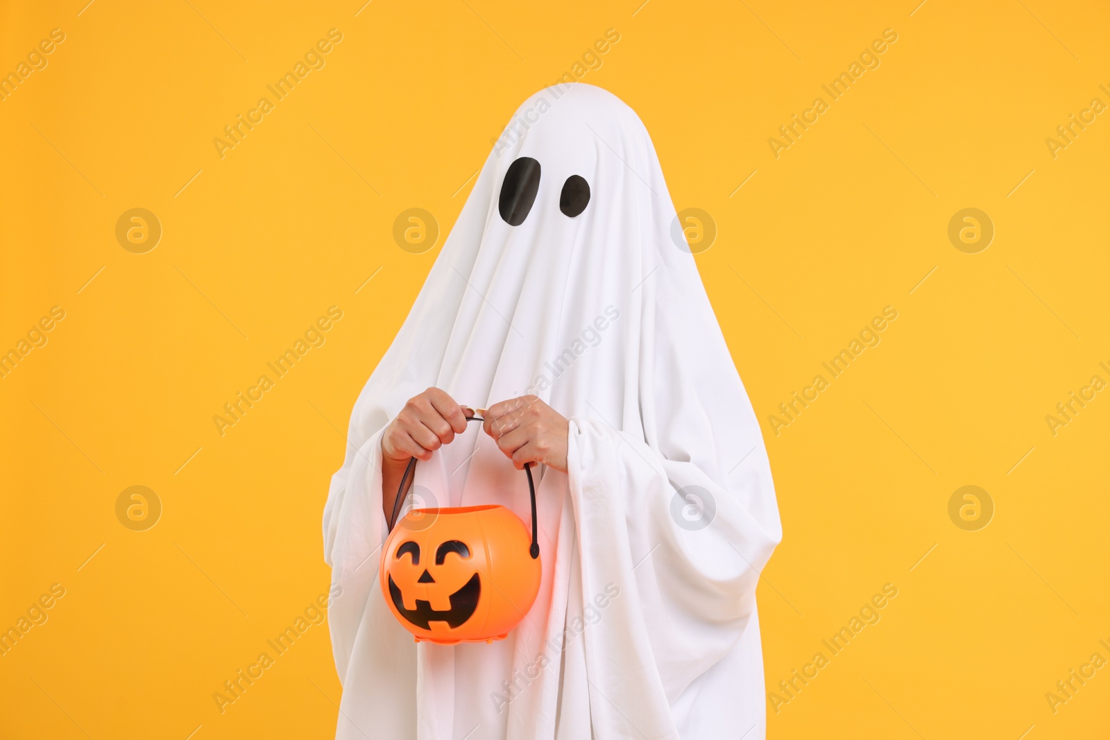 Photo of Woman in white ghost costume holding pumpkin bucket on yellow background. Halloween celebration