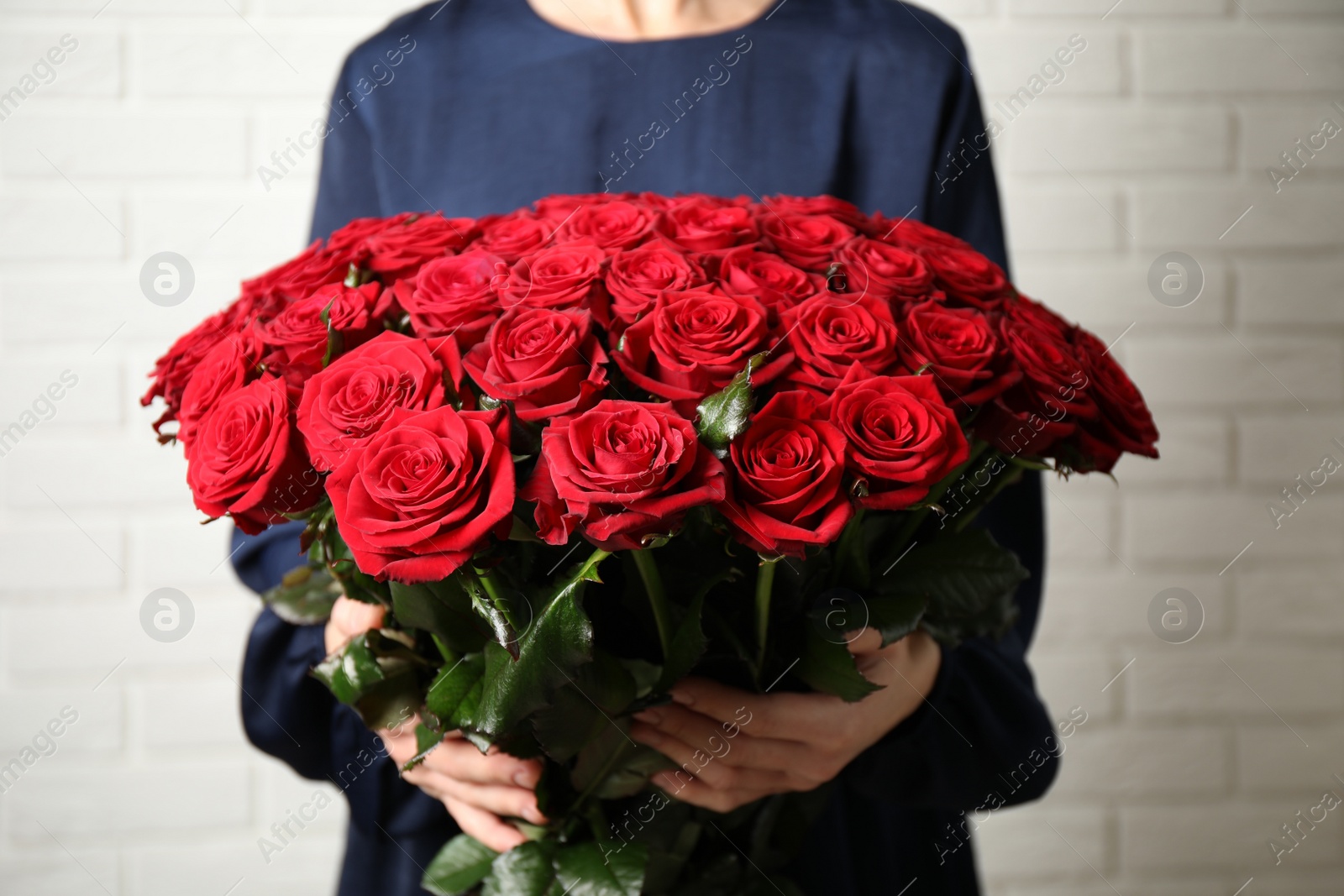 Photo of Woman holding luxury bouquet of fresh red roses near white brick wall, closeup