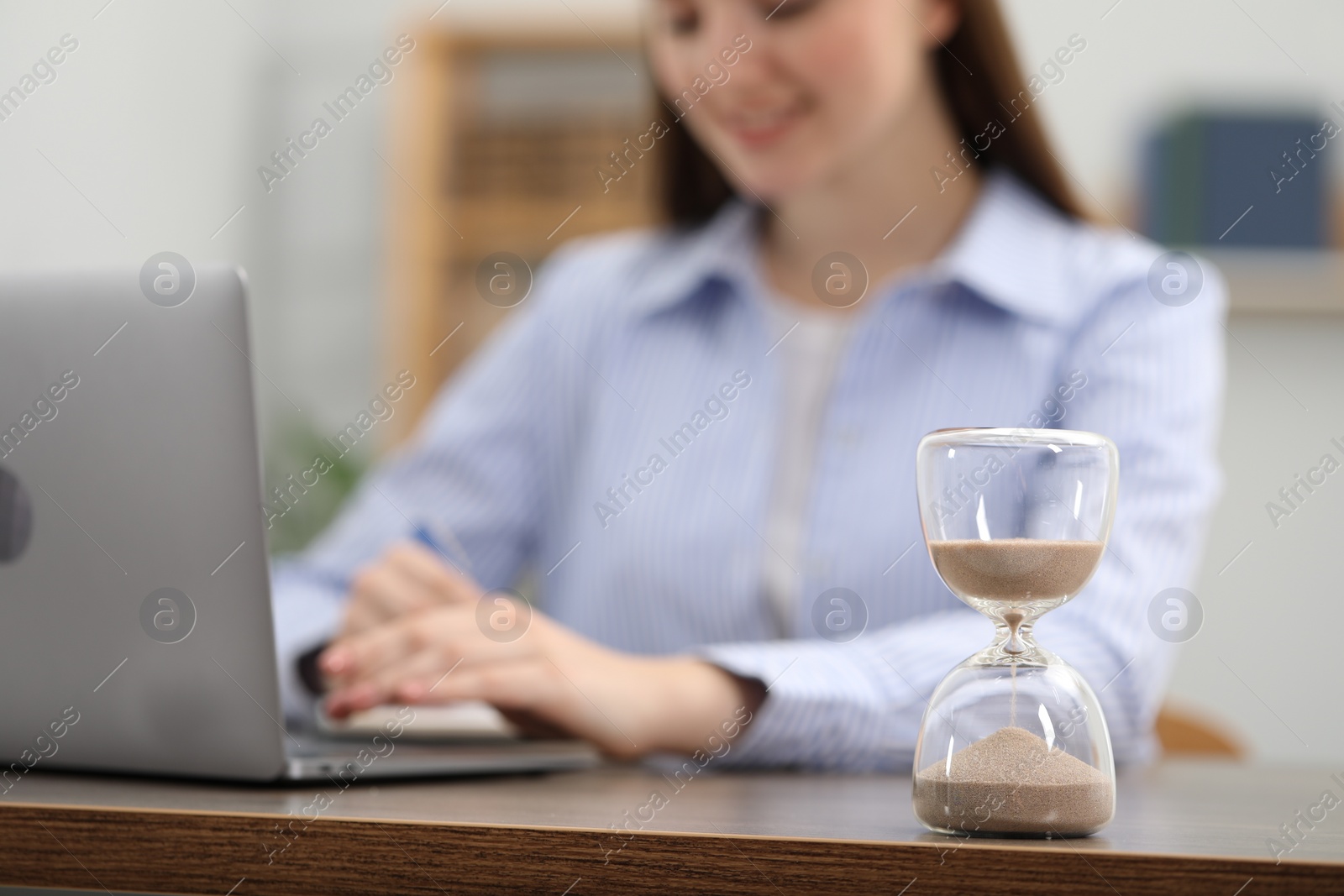 Photo of Hourglass with flowing sand on desk. Woman using laptop indoors, selective focus