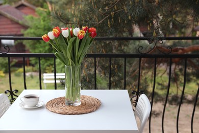 Photo of Beautiful colorful tulips in glass vase and cup of drink on white table at balcony, space for text