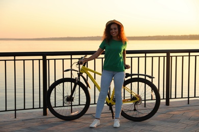 Beautiful young African-American woman with bicycle on city waterfront at sunset