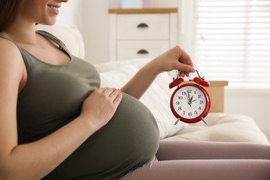 Photo of Young pregnant woman holding alarm clock near her belly at home, closeup. Time to give birth