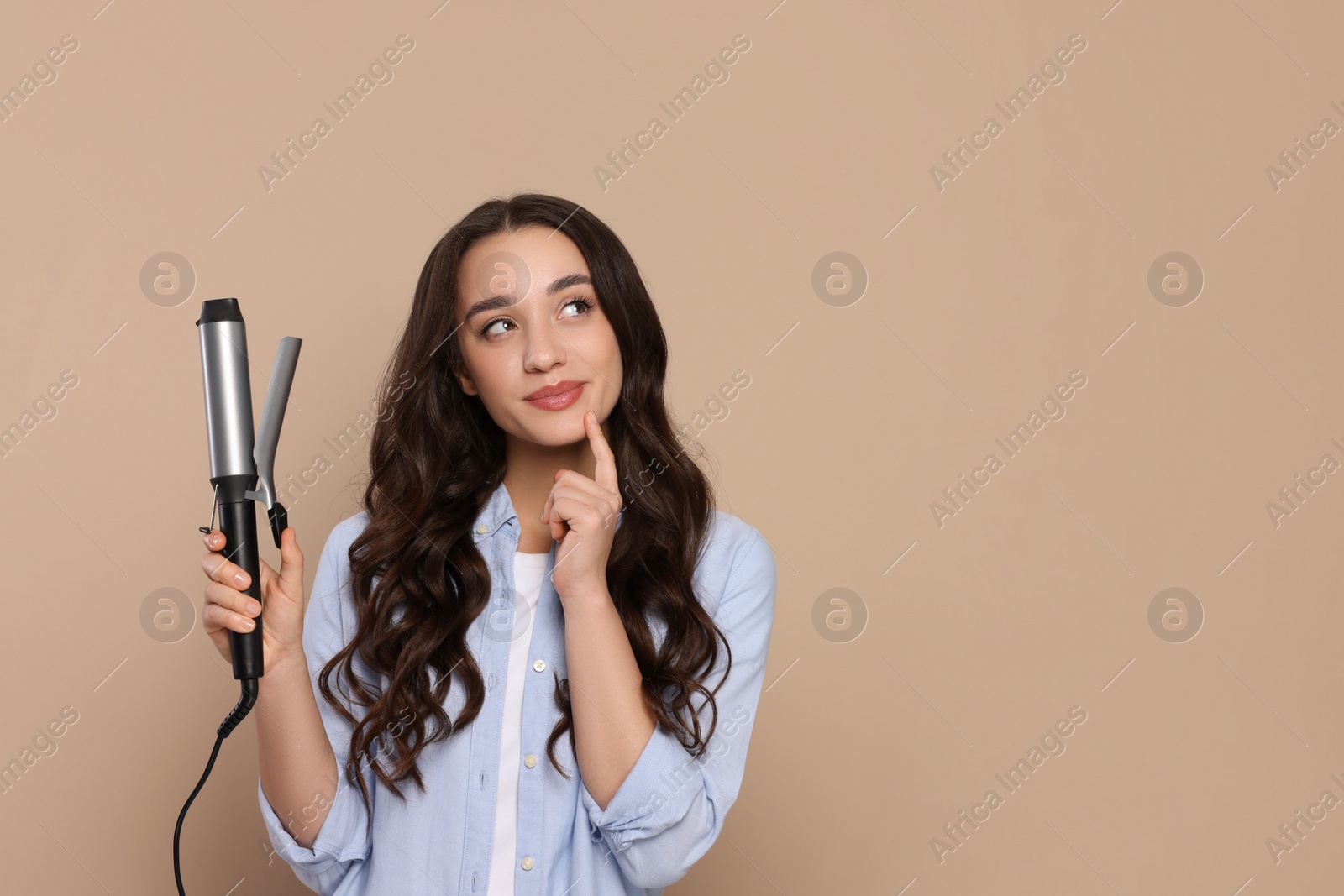 Photo of Thoughtful woman with curling hair iron on beige background. Space for text