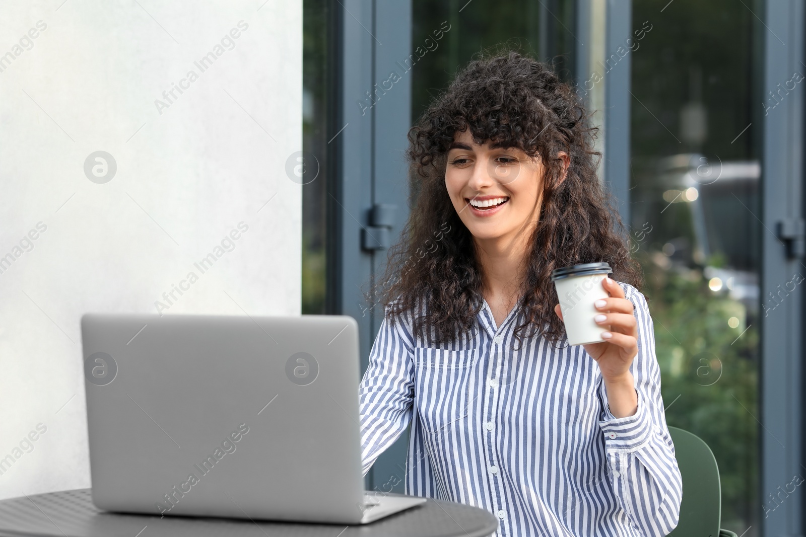 Photo of Happy young woman with cup of coffee using modern laptop at table outdoors