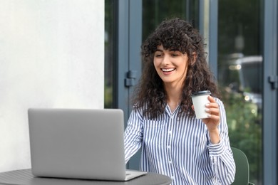 Happy young woman with cup of coffee using modern laptop at table outdoors