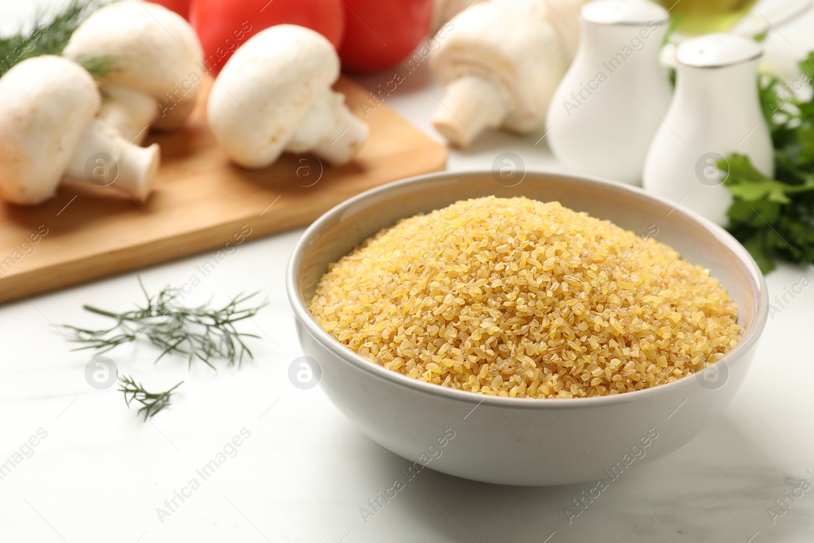Photo of Raw bulgur in bowl, vegetables and spices on white table, closeup