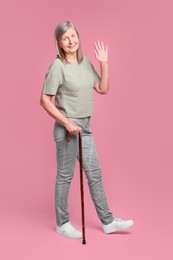 Senior woman with walking cane waving on pink background