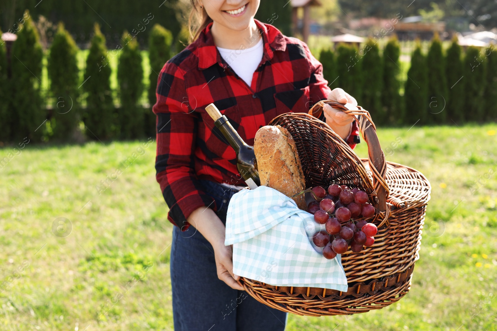 Photo of Woman holding wicker basket with wine and snacks in park, closeup. Picnic season