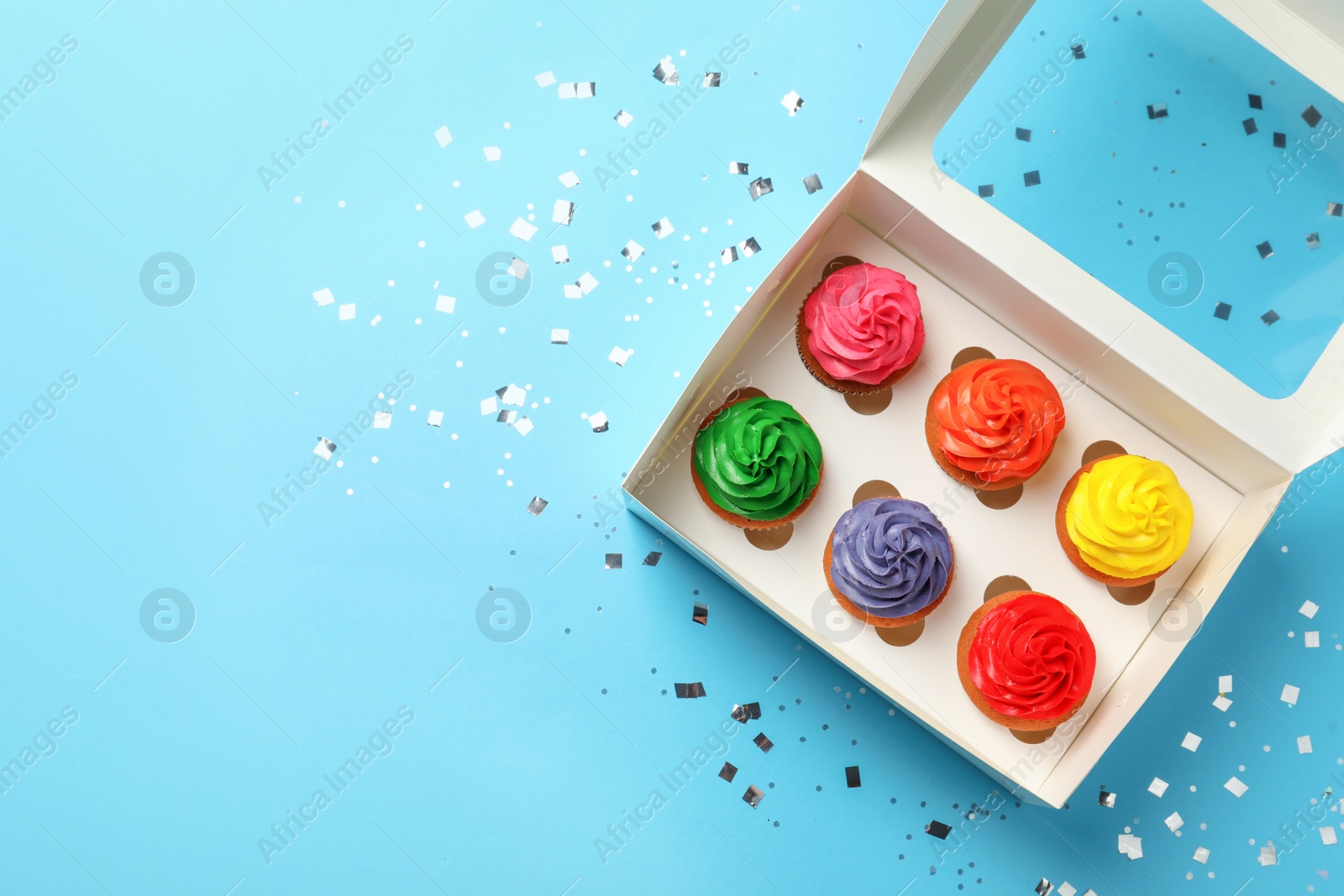 Photo of Box with delicious colorful cupcakes and confetti on light blue background, flat lay. Space for text