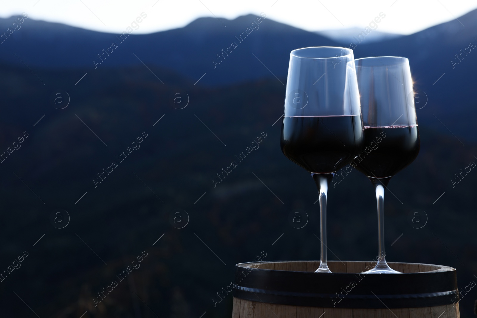 Photo of Glasses of red wine on barrel against mountain landscape, space for text