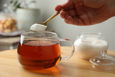 Woman adding sugar into aromatic tea at wooden table indoors, closeup