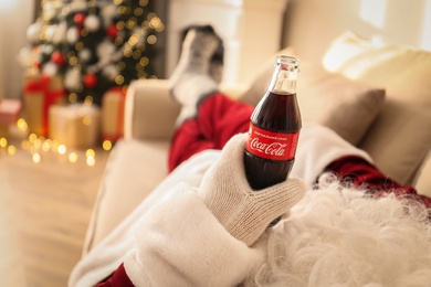 Photo of MYKOLAIV, UKRAINE - JANUARY 18, 2021: Santa Claus with Coca-Cola bottle resting on sofa in room, closeup