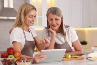 Photo of Happy mature mother and her daughter using tablet while cooking in kitchen