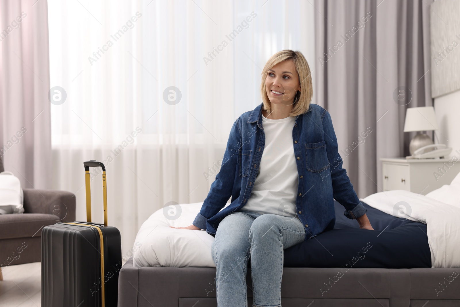 Photo of Smiling guest with suitcase relaxing on bed in stylish hotel room