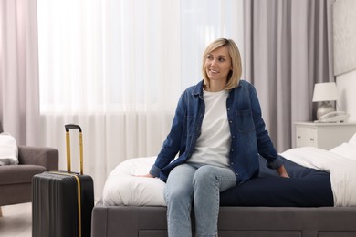 Smiling guest with suitcase relaxing on bed in stylish hotel room