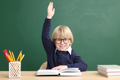 Photo of Happy little school child raising hand while sitting at desk with books near chalkboard