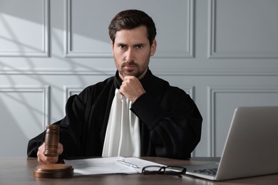 Judge with gavel, papers and laptop sitting at wooden table indoors