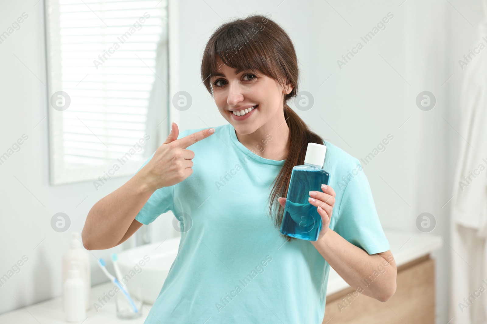 Photo of Young woman with mouthwash pointing at her healthy teeth in bathroom