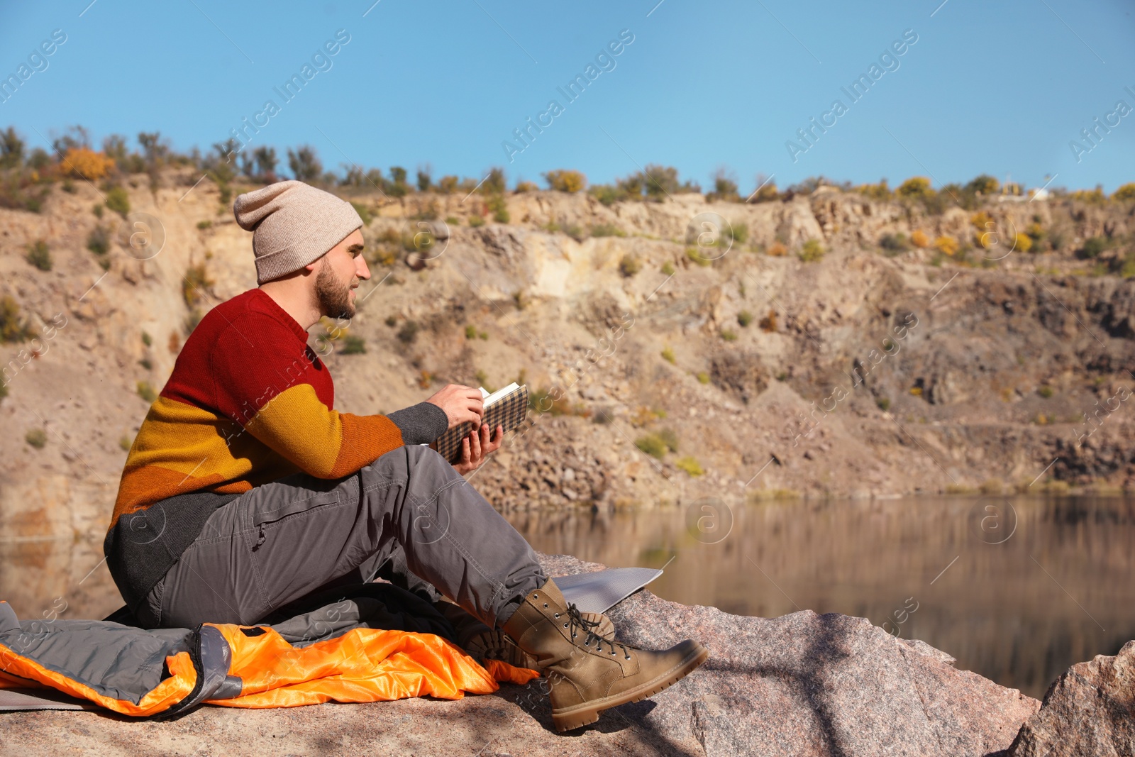 Photo of Male camper reading book while sitting on sleeping bag in wilderness. Space for text