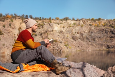 Photo of Male camper reading book while sitting on sleeping bag in wilderness. Space for text