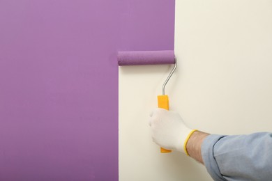 Photo of Man applying violet paint with roller brush on white wall, closeup