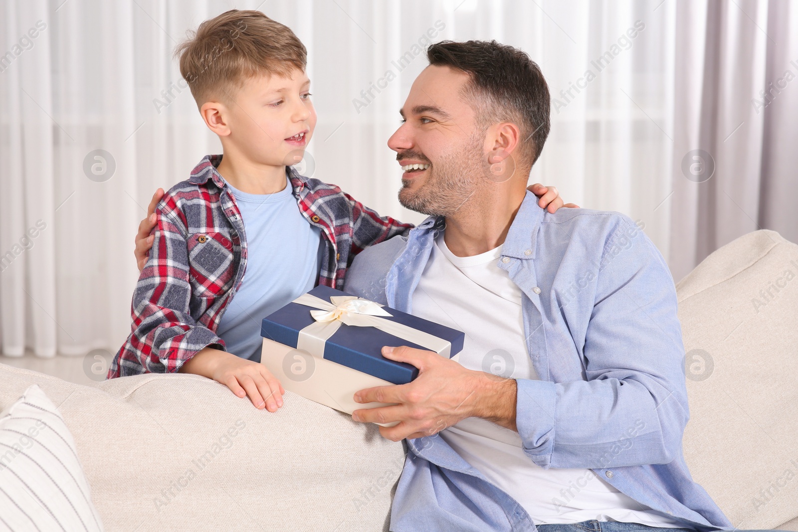 Photo of Cute little boy presenting his father with gift on sofa at home