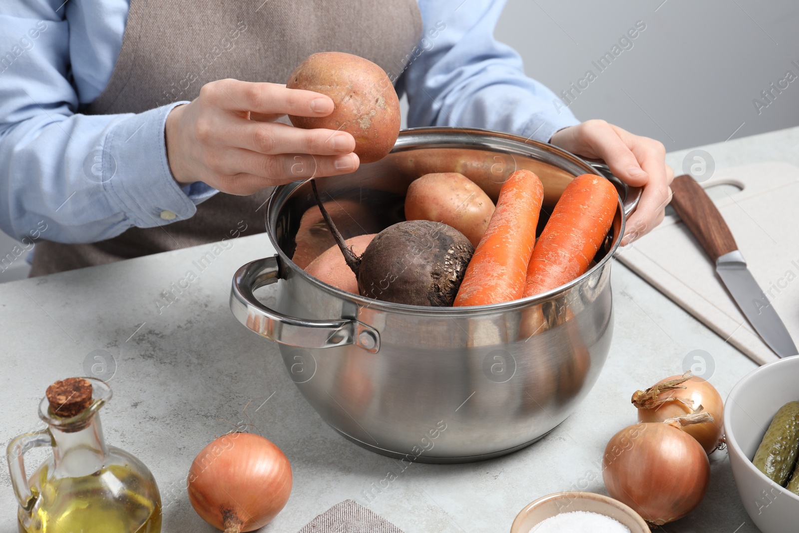 Photo of Woman putting potato into pot with fresh vegetables at white table, closeup. Cooking vinaigrette salad