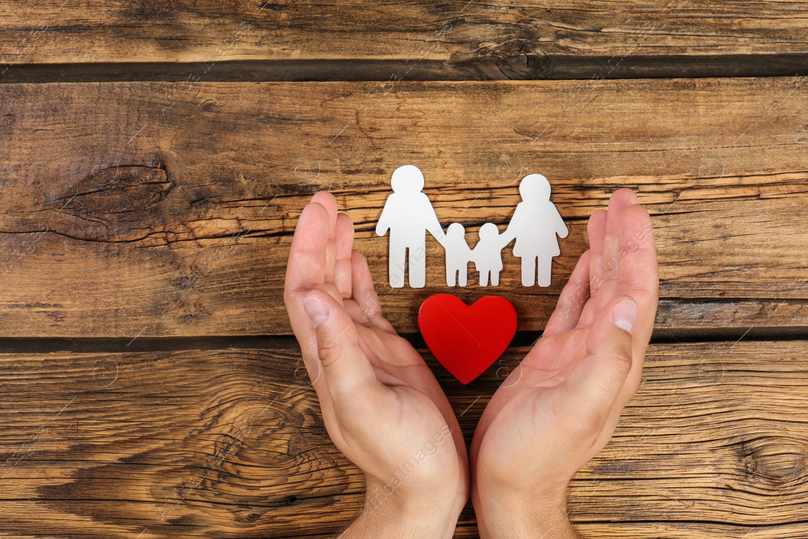 Photo of Young man protecting family figure and red heart with his hands on wooden background, top view. Space for text