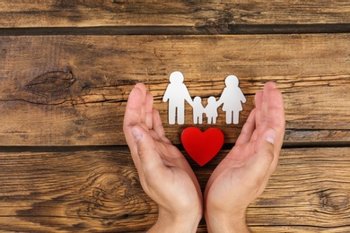 Photo of Young man protecting family figure and red heart with his hands on wooden background, top view. Space for text