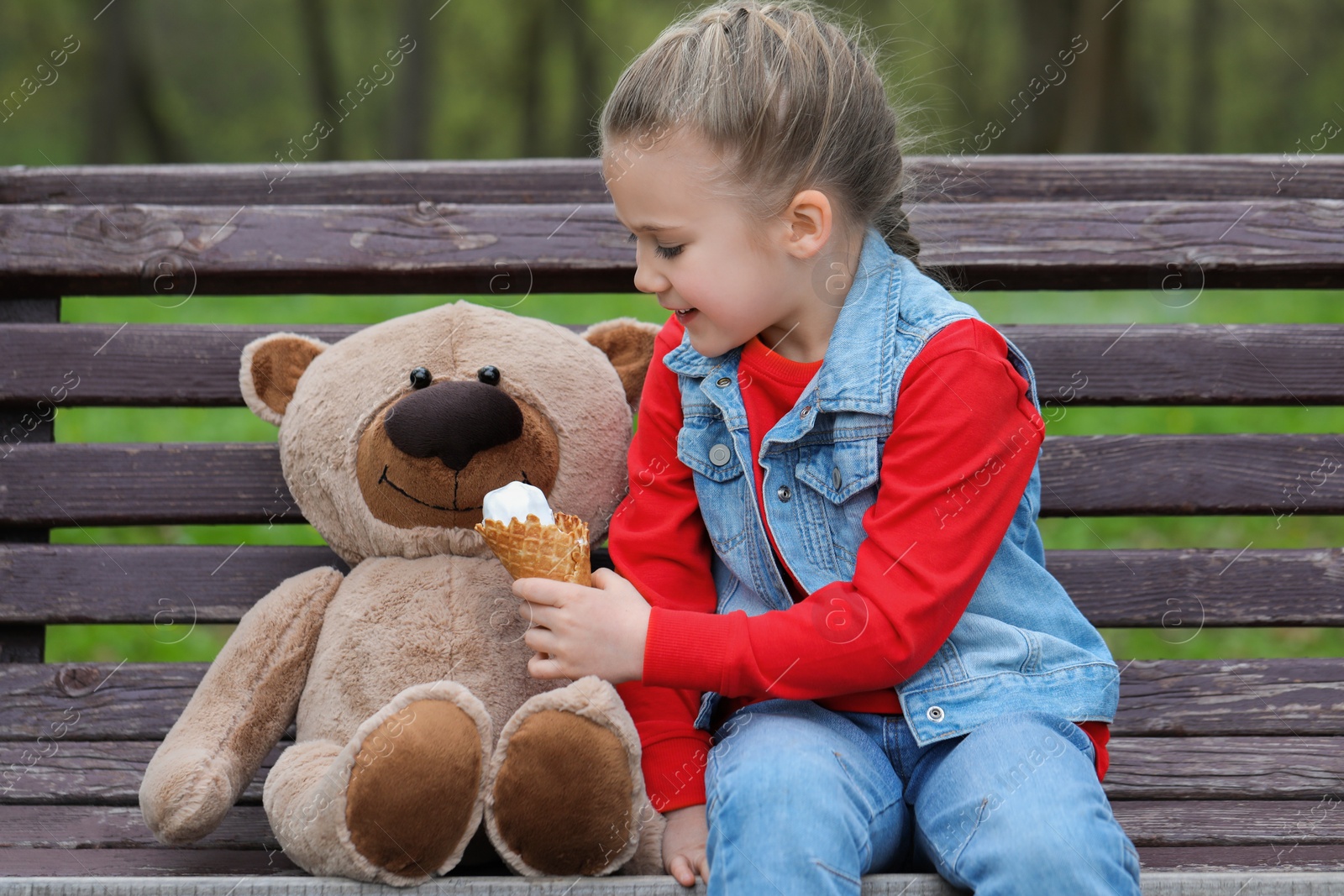 Photo of Little girl with teddy bear and ice cream on wooden bench outdoors